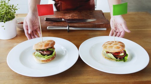 Två vita tallrikar med varsin hamburgare med bröd på. Båda likadana med sallad och grönsaker som sticker ut men ingredienserna i burgarna är olika. Den med kött har en röd flagga på träpinne instinkt i sig och den veganska en grön flagga.