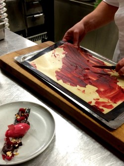 Halloncrisp petas bort med kniv från bakplåtspapper på plåt. Framför står en tallrik med chokladcreme och hallonsorbé som ska garneras blir crispen