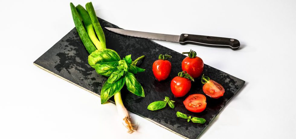Basilika och tomater på en rektangulär svart bräda, en kniv ligger ovanför.