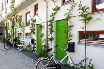 Husbyggnad, vit fasad med gröna dörrar, klängväxter växer upp längs väggen vid sidorna av de båda dörrarna, vit cykel parkerad framför den ena i förgrunden. 