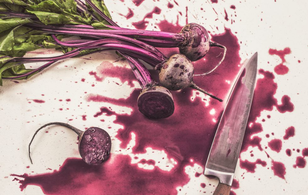 Delade rödbetor med blast på vit bänk, kniv till höger och rödbetssaft stänkt runt om