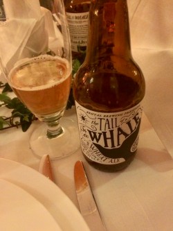 Brun ölflaska till höger om ölglas med lite öl intill tallrik och bestick på vit linneduk