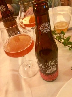 Brun glasflaska till höger om ett ölglas med lite rosafärgad öl i. Står på vit linneduk