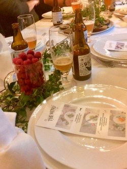 Brun ölflaska till höger om ölglas med lite öl intill tallrik och bestick på vit linneduk. Bredvid stort ölglas fyllt med färska hallon, diukningen dekorerad med gröna blad