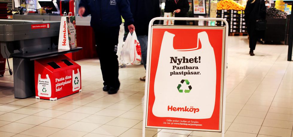 En golvskylt vid butikskassa, på skylten en målad plastkasse i vitt med texten "Nyhet! Pantbara plastpåsar" i svart