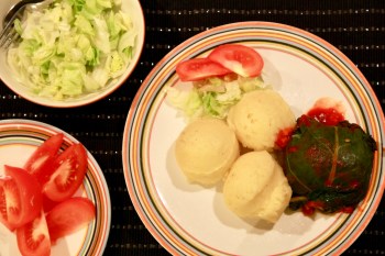 En tallrik med tre kulor potatismos, en mangolddolm och lite sallad och tomat. Till vänster fat med klyftade tomater och en skål med grönsallad.