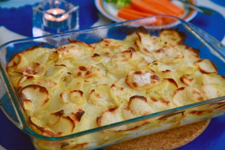 Potatisgratäng i en rektangulär glasform, i bakgrunden ett tänt ljus och ett fat med morotsstavar
