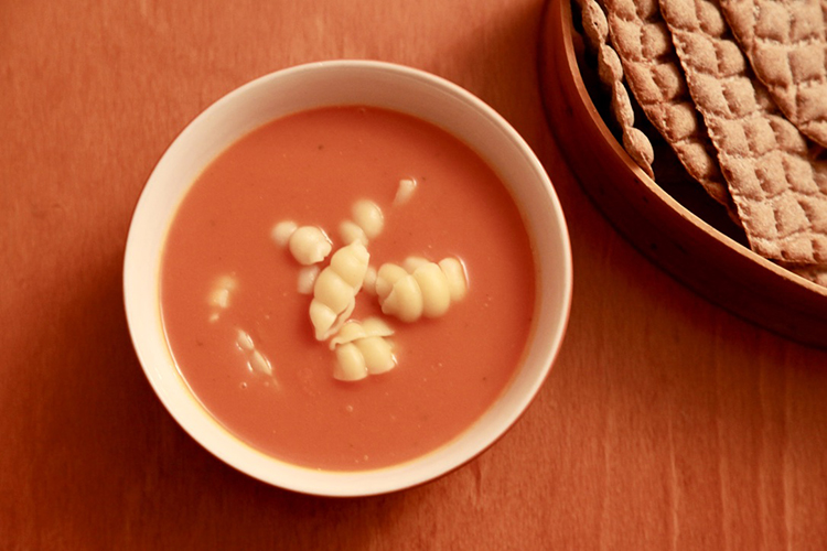 En skål med tomatsoppa toppad med pastasnäckor sedd uppifrån. Till höger en brödform med hårt bröd.