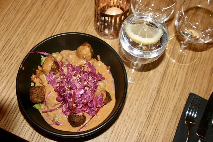 Svart skål med kikärtsbullar, picklade rödlök och jordnötssås sett uppifrån, ovanför glas med vetten och citronskiva samt ett tänt värmeljus