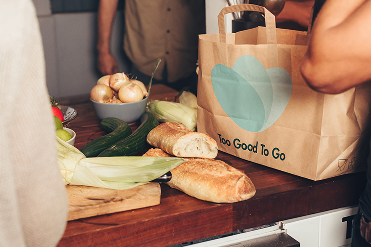 En bänk med bröd och grönsaker, bredvid en pappkasse med loggan Too Good To Go