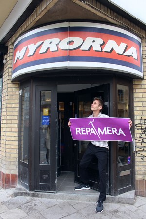 En man håller i en lila skylt med texten REPAMERA i vitt. Står i en dörröppning, ovanför hänger skylt med Myrornas logo på