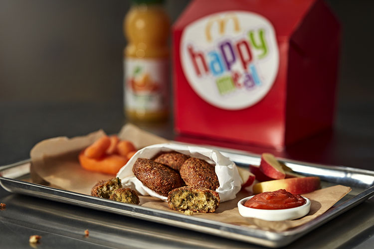 En bricka med falafel, skål med ketchup och morötter, i bakgrunden förpackning till Happy Meal