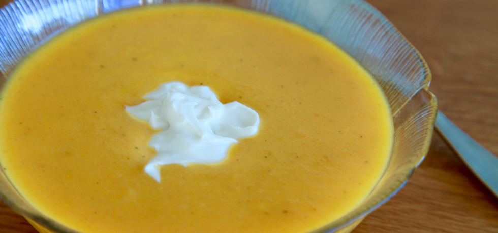 Gul morotssoppa i glasskål med sked till höger sett uppifrån