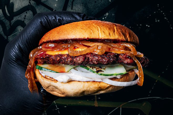 En hand med svart handske håller en burgare med bröd och tillbehör